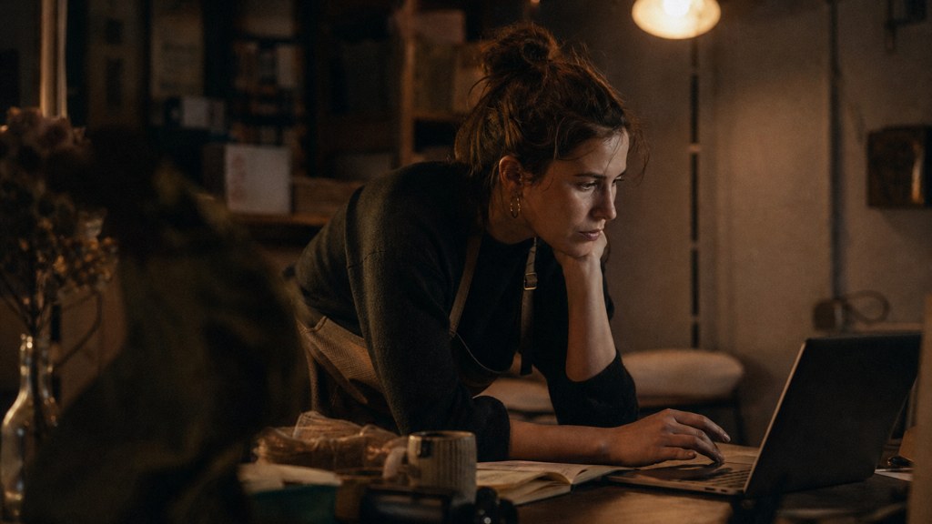 A small-business owner in an apron leaning over a laptop on a cluttered back-office desk late in the day, photographed in Cardiff