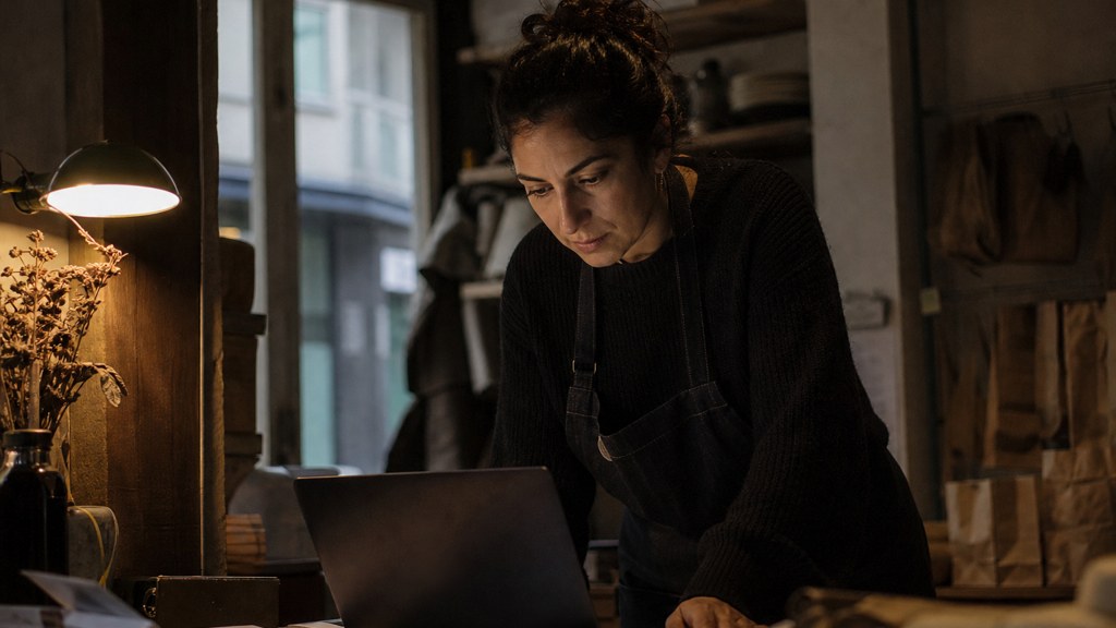 A small-business owner in an apron leaning over a laptop on a cluttered back-office desk late in the day, photographed in Cardiff