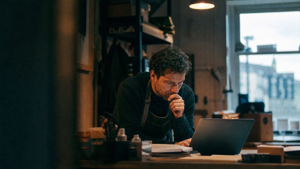 A small-business owner in an apron leaning over a laptop on a cluttered back-office desk late in the day, photographed in Cardiff