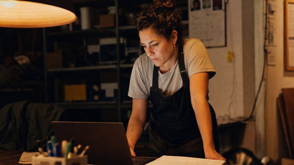 A small-business owner in an apron leaning over a laptop on a cluttered back-office desk late in the day, photographed in Chandler