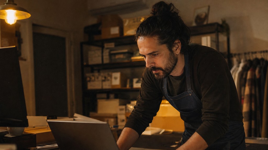 A small-business owner in an apron leaning over a laptop on a cluttered back-office desk late in the day, photographed in Chandler