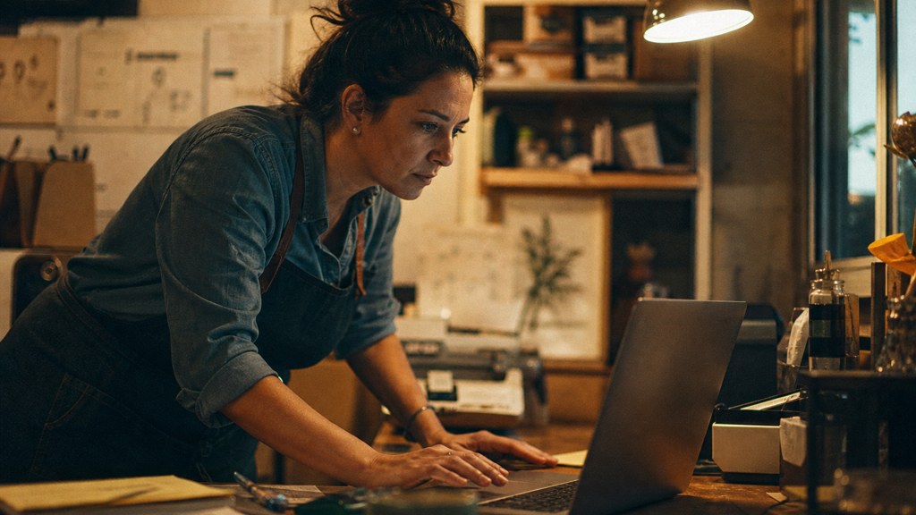 A small-business owner in an apron leaning over a laptop on a cluttered back-office desk late in the day, photographed in Chandler
