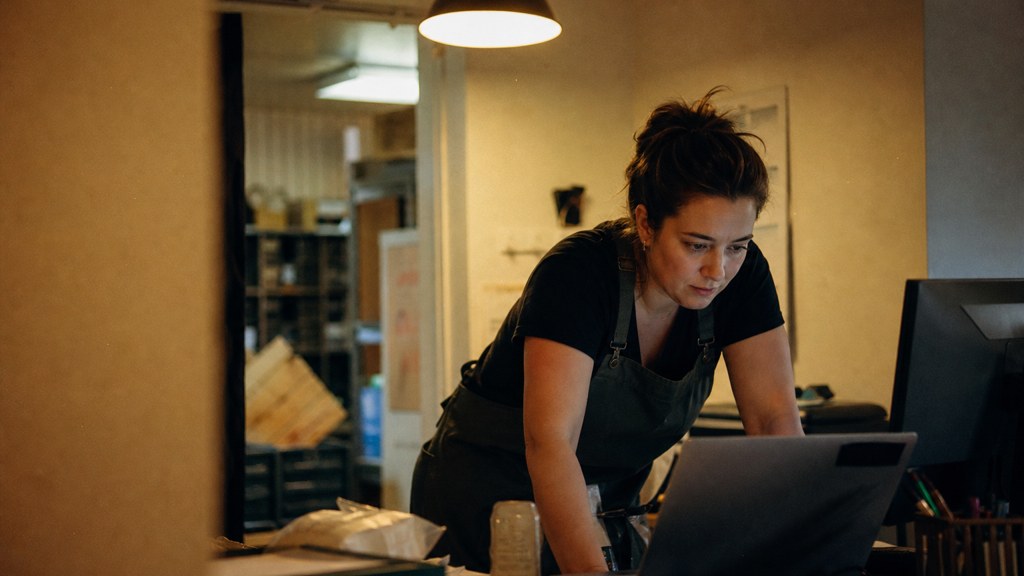 A small-business owner in an apron leaning over a laptop on a cluttered back-office desk late in the day, photographed in Chandler