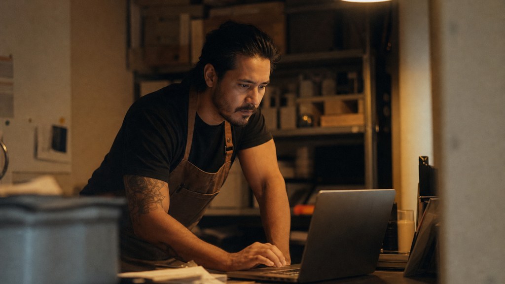 A small-business owner in an apron leaning over a laptop on a cluttered back-office desk late in the day, photographed in Chandler