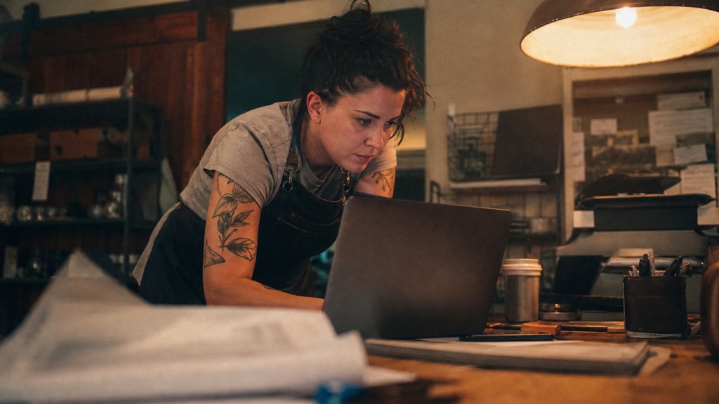A small-business owner in an apron leaning over a laptop on a cluttered back-office desk late in the day, photographed in Chandler
