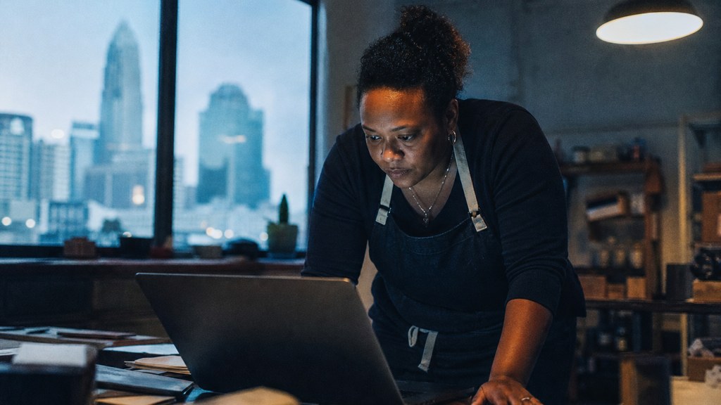 A small-business owner in an apron leaning over a laptop on a cluttered back-office desk late in the day, photographed in Charlotte