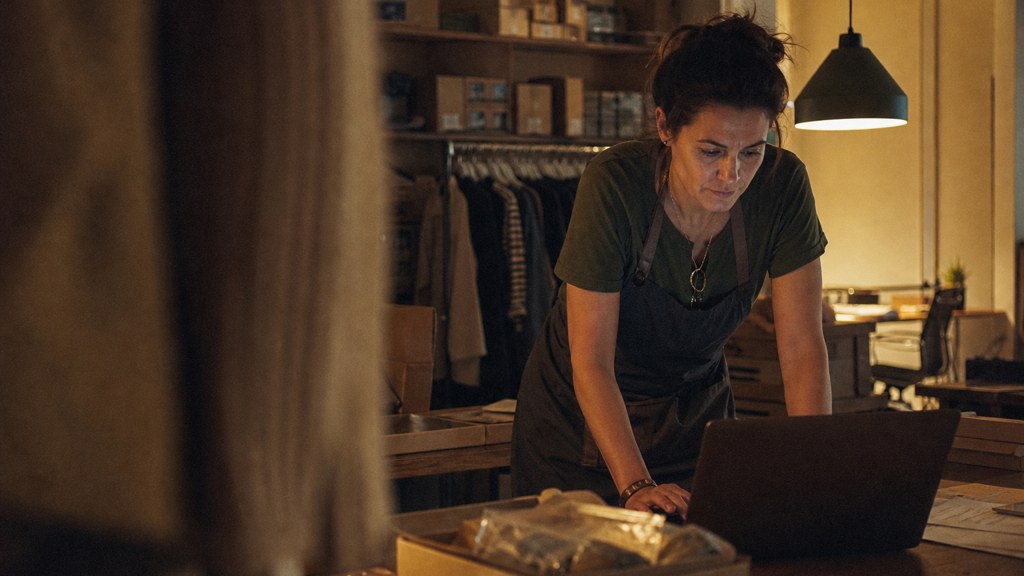 A small-business owner in an apron leaning over a laptop on a cluttered back-office desk late in the day, photographed in Charlotte
