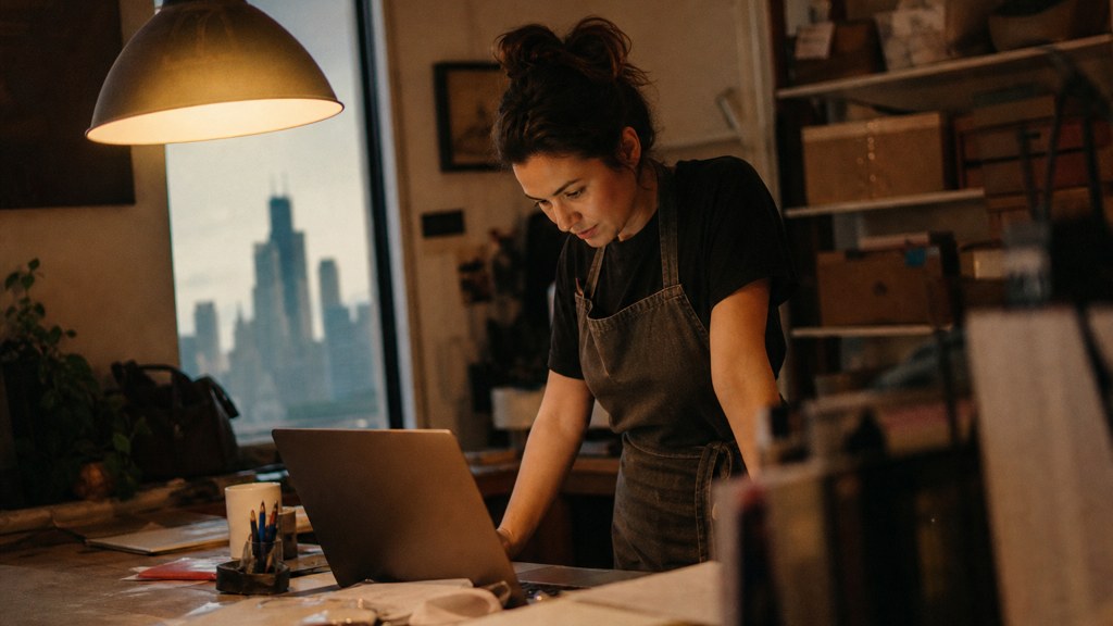 A small-business owner in an apron leaning over a laptop on a cluttered back-office desk late in the day, photographed in Chicago