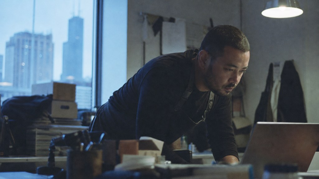 A small-business owner in an apron leaning over a laptop on a cluttered back-office desk late in the day, photographed in Chicago