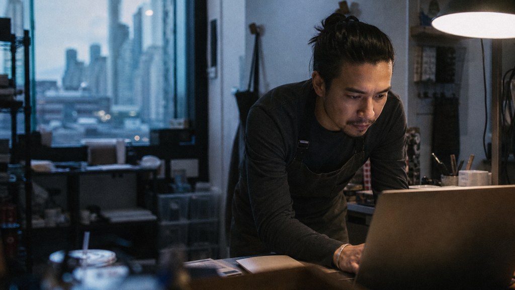 A small-business owner in an apron leaning over a laptop on a cluttered back-office desk late in the day, photographed in Chicago