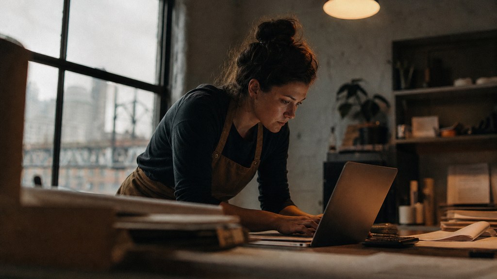 A small-business owner in an apron leaning over a laptop on a cluttered back-office desk late in the day, photographed in Cincinnati