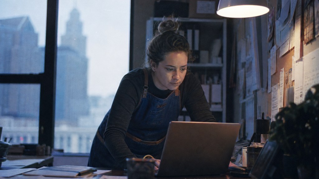 A small-business owner in an apron leaning over a laptop on a cluttered back-office desk late in the day, photographed in Cleveland