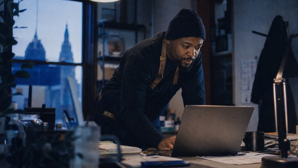 A small-business owner in an apron leaning over a laptop on a cluttered back-office desk late in the day, photographed in Cleveland