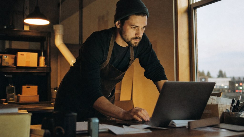 A small-business owner in an apron leaning over a laptop on a cluttered back-office desk late in the day, photographed in Colorado Springs