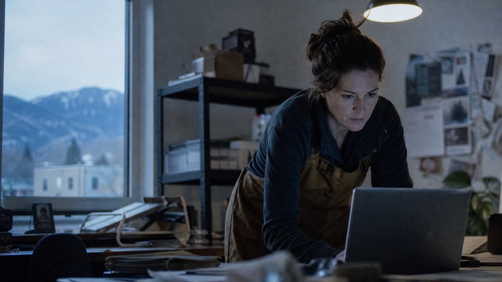 A small-business owner in an apron leaning over a laptop on a cluttered back-office desk late in the day, photographed in Colorado Springs