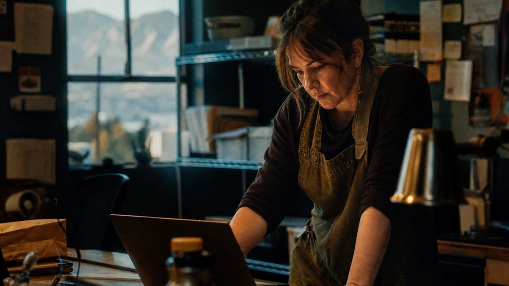 A small-business owner in an apron leaning over a laptop on a cluttered back-office desk late in the day, photographed in Colorado Springs