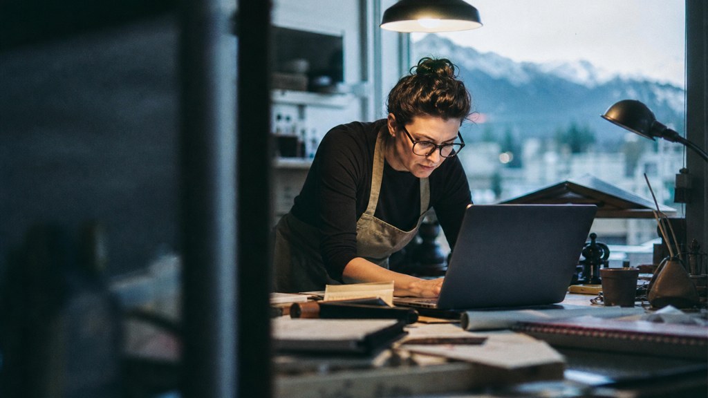 A small-business owner in an apron leaning over a laptop on a cluttered back-office desk late in the day, photographed in Colorado Springs