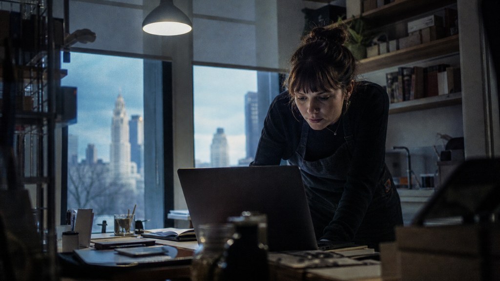 A small-business owner in an apron leaning over a laptop on a cluttered back-office desk late in the day, photographed in Columbus