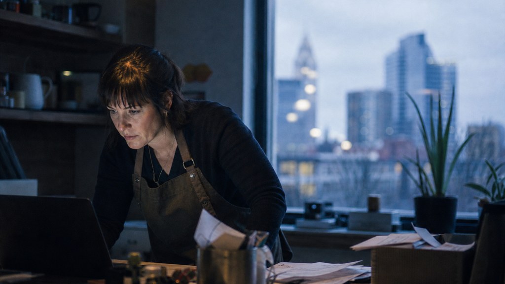 A small-business owner in an apron leaning over a laptop on a cluttered back-office desk late in the day, photographed in Columbus
