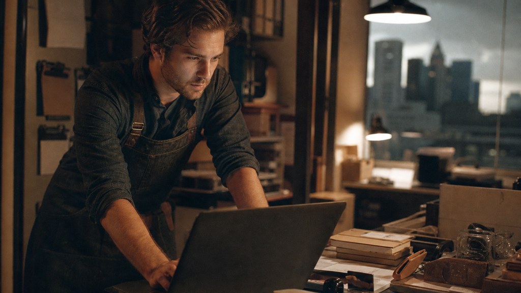 A small-business owner in an apron leaning over a laptop on a cluttered back-office desk late in the day, photographed in Columbus