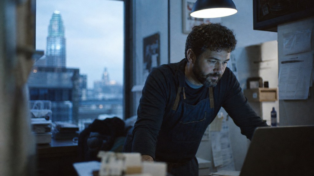 A small-business owner in an apron leaning over a laptop on a cluttered back-office desk late in the day, photographed in Columbus