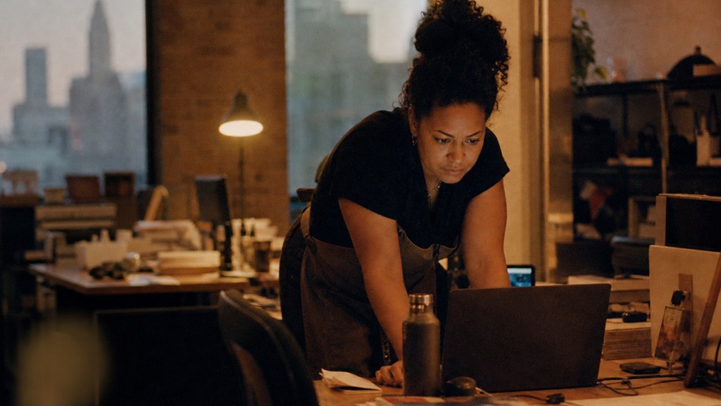 A small-business owner in an apron leaning over a laptop on a cluttered back-office desk late in the day, photographed in Columbus