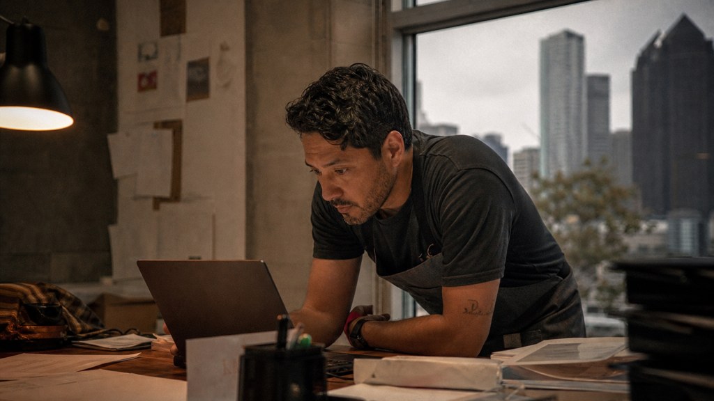 A small-business owner in an apron leaning over a laptop on a cluttered back-office desk late in the day, photographed in Dallas