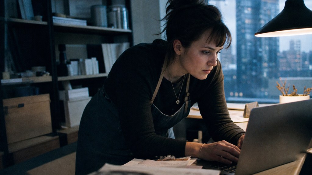 A small-business owner in an apron leaning over a laptop on a cluttered back-office desk late in the day, photographed in Denver