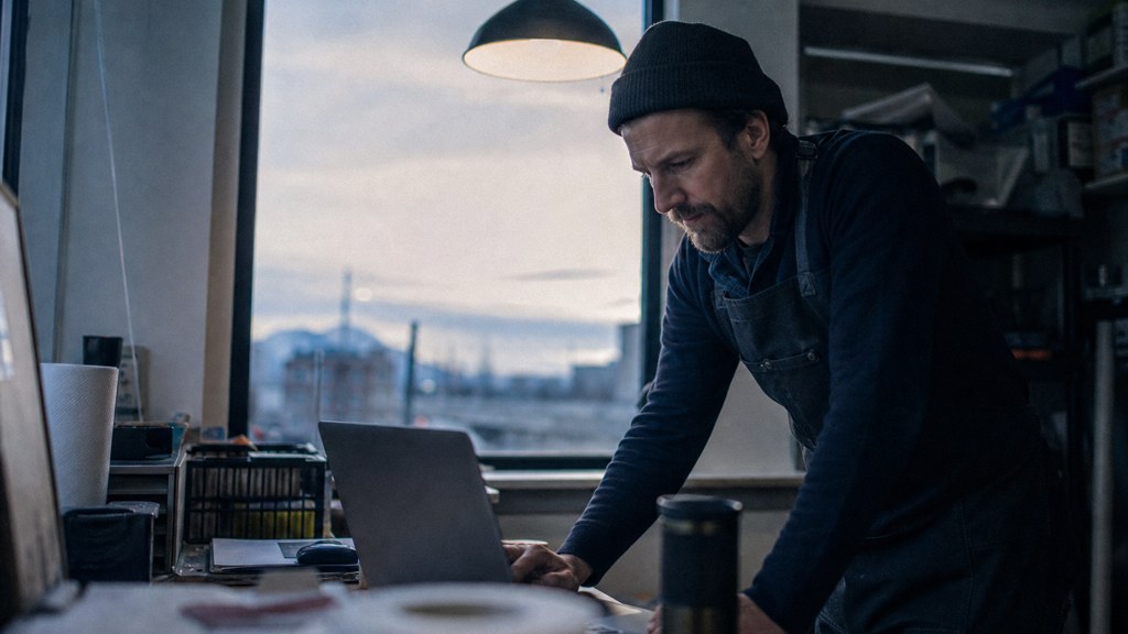 A small-business owner in an apron leaning over a laptop on a cluttered back-office desk late in the day, photographed in Denver