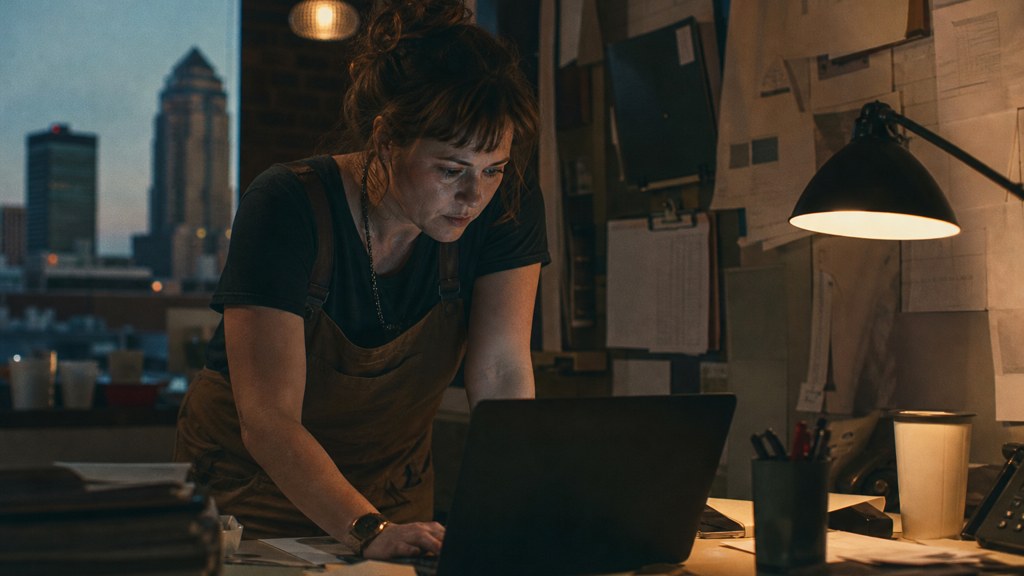 A small-business owner in an apron leaning over a laptop on a cluttered back-office desk late in the day, photographed in Des Moines