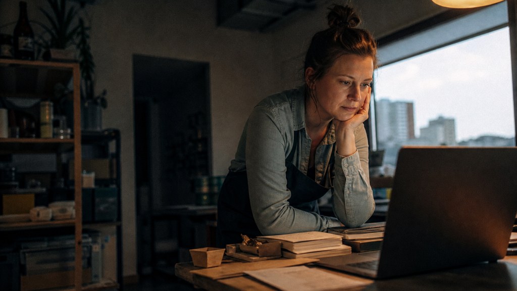 A small-business owner in an apron leaning over a laptop on a cluttered back-office desk late in the day, photographed in Des Moines