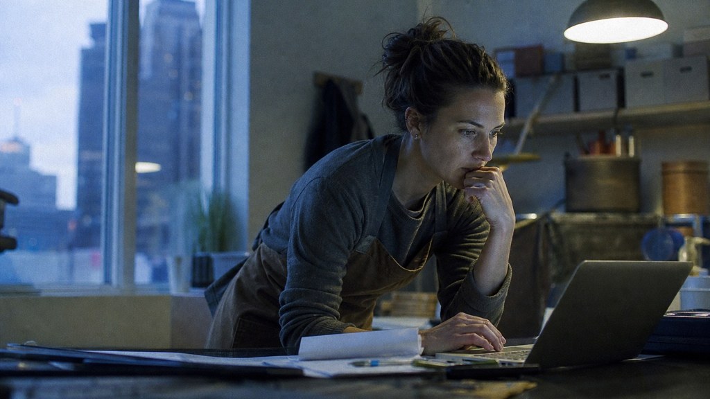 A small-business owner in an apron leaning over a laptop on a cluttered back-office desk late in the day, photographed in Des Moines