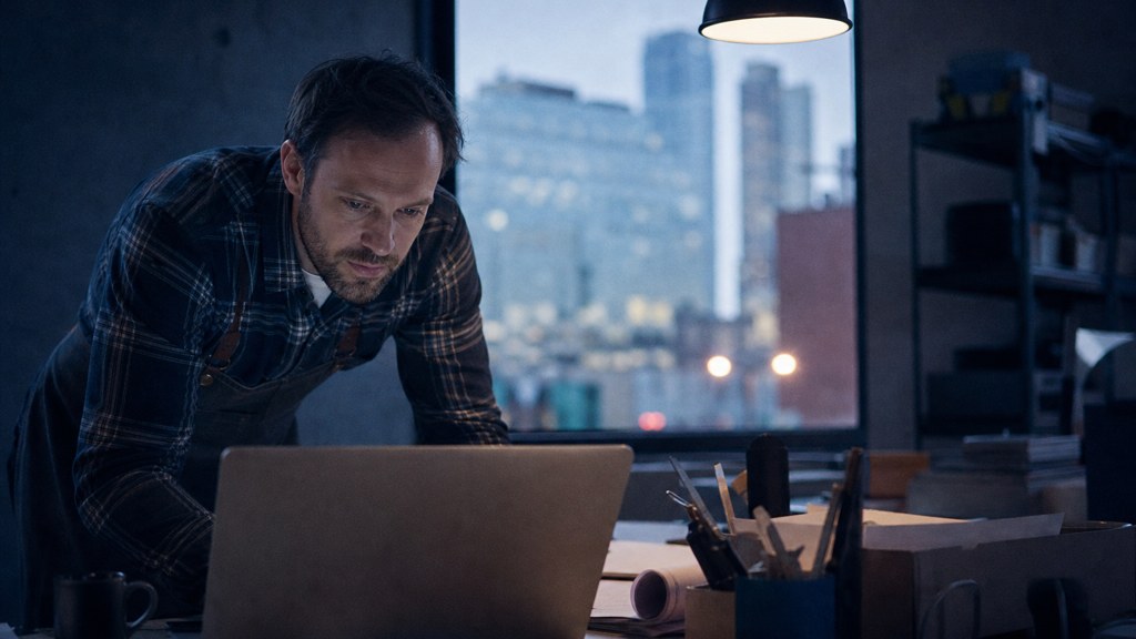 A small-business owner in an apron leaning over a laptop on a cluttered back-office desk late in the day, photographed in Des Moines