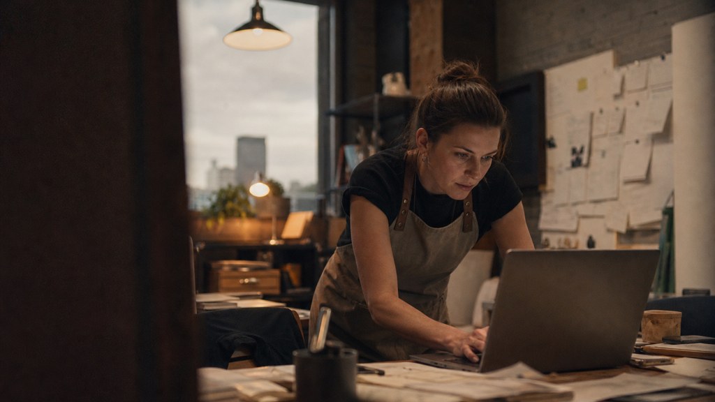 A small-business owner in an apron leaning over a laptop on a cluttered back-office desk late in the day, photographed in Des Moines