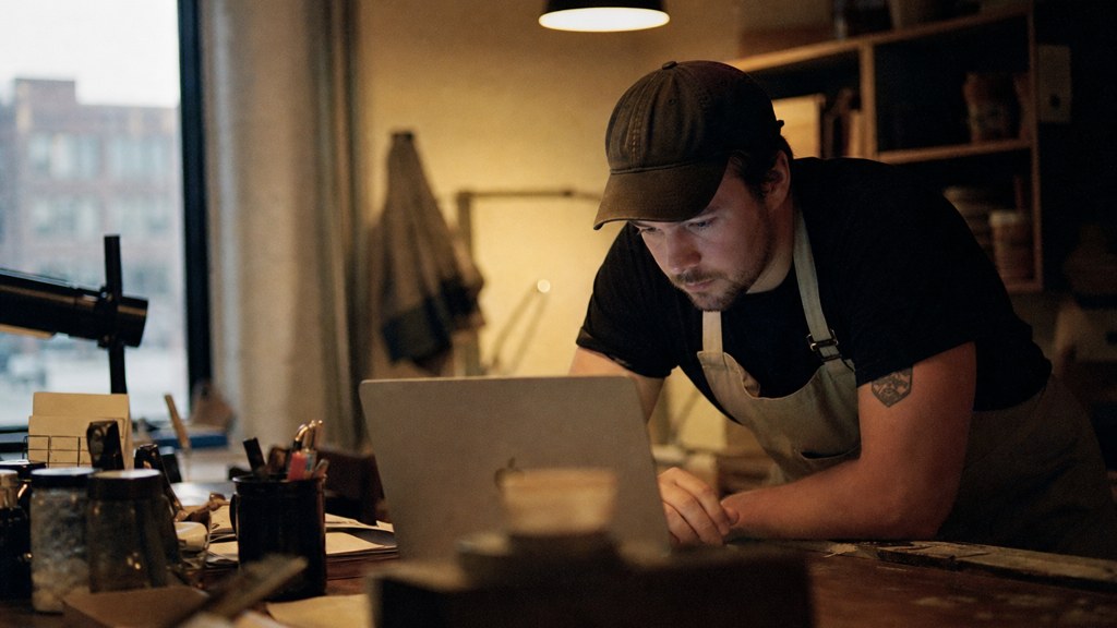 A small-business owner in an apron leaning over a laptop on a cluttered back-office desk late in the day, photographed in Des Moines