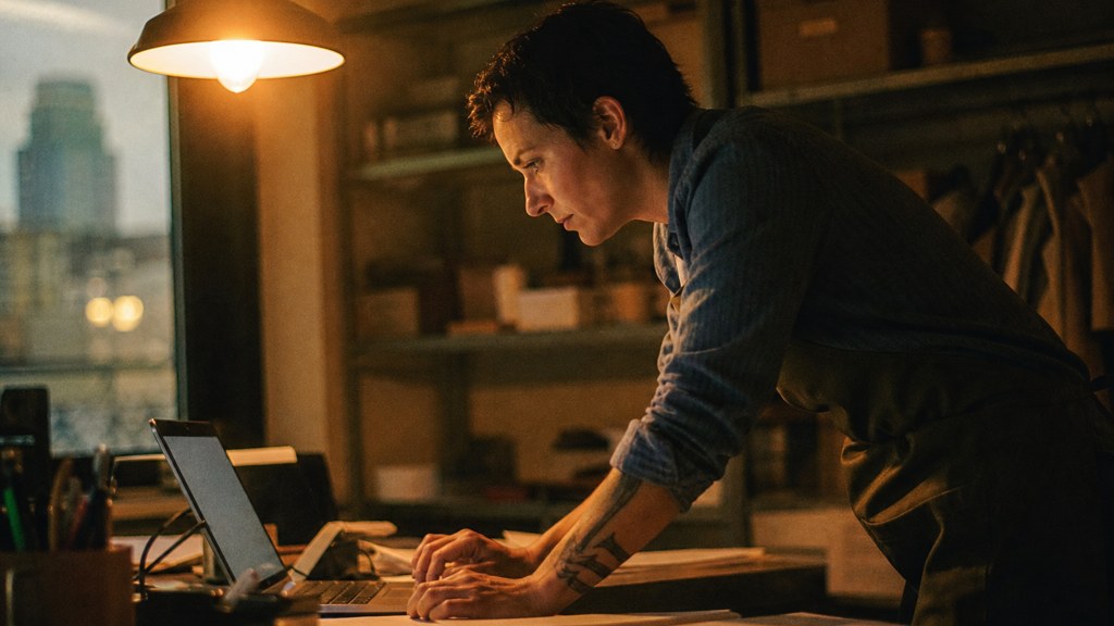 A small-business owner in an apron leaning over a laptop on a cluttered back-office desk late in the day, photographed in Des Moines