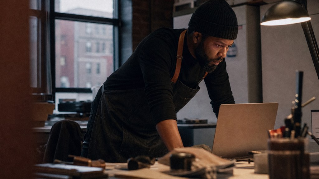 A small-business owner in an apron leaning over a laptop on a cluttered back-office desk late in the day, photographed in Detroit