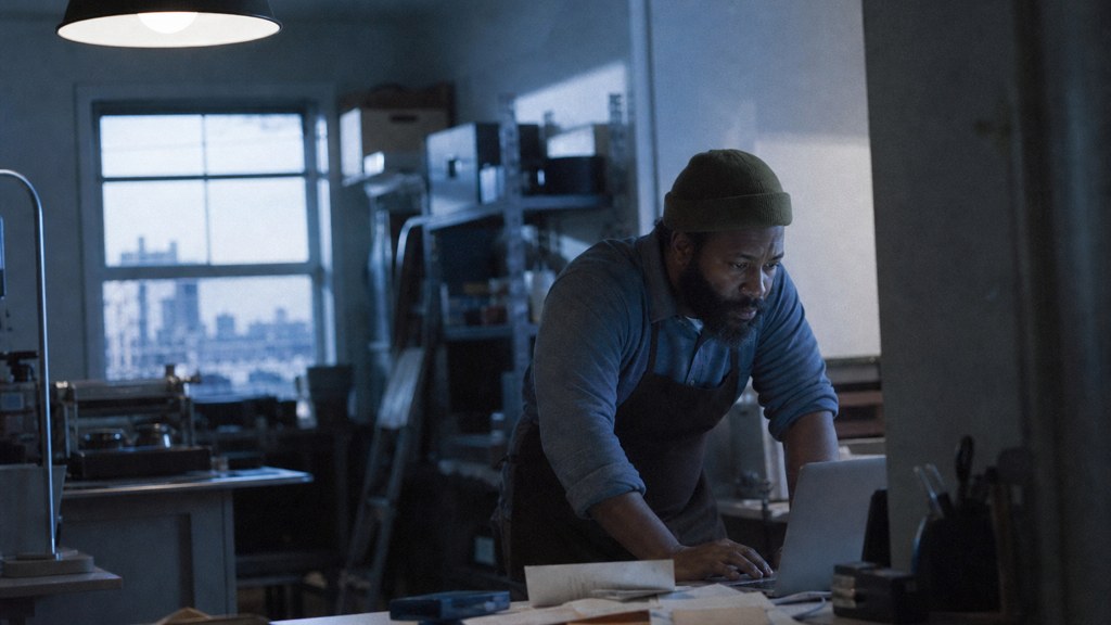 A small-business owner in an apron leaning over a laptop on a cluttered back-office desk late in the day, photographed in Detroit