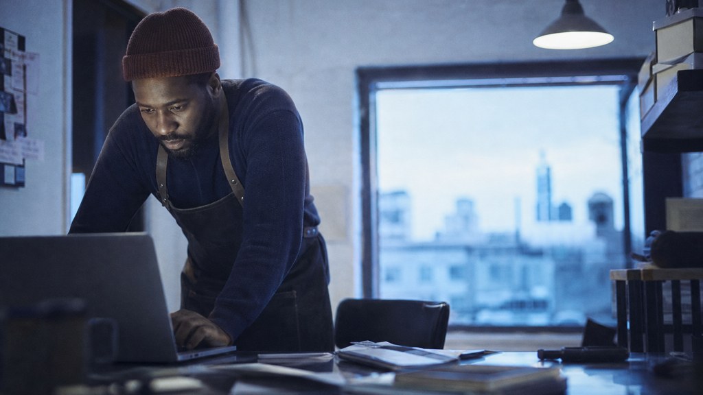 A small-business owner in an apron leaning over a laptop on a cluttered back-office desk late in the day, photographed in Detroit