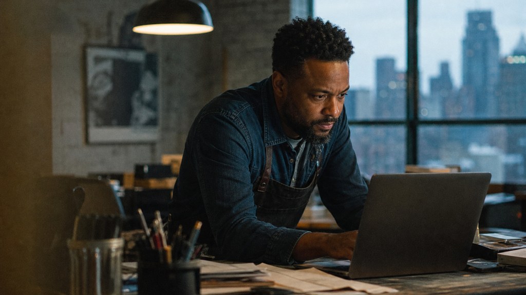 A small-business owner in an apron leaning over a laptop on a cluttered back-office desk late in the day, photographed in Detroit