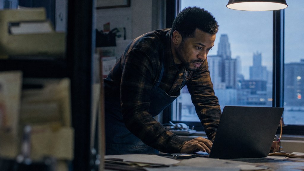 A small-business owner in an apron leaning over a laptop on a cluttered back-office desk late in the day, photographed in Detroit