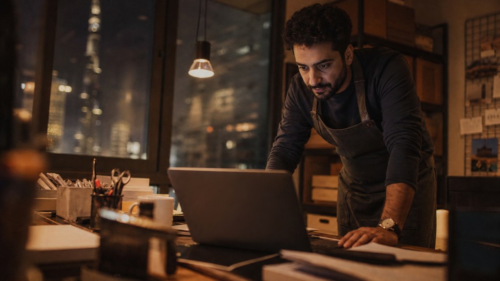 A small-business owner in an apron leaning over a laptop on a cluttered back-office desk late in the day, photographed in Dubai
