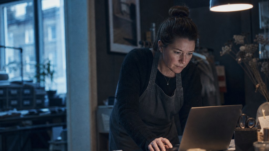 A small-business owner in an apron leaning over a laptop on a cluttered back-office desk late in the day, photographed in Dublin