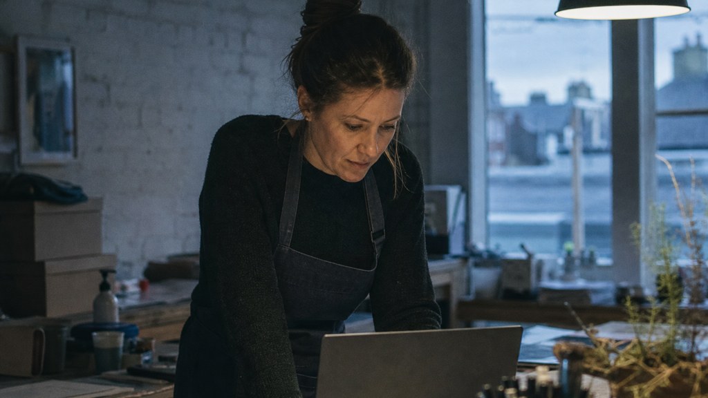 A small-business owner in an apron leaning over a laptop on a cluttered back-office desk late in the day, photographed in Dublin