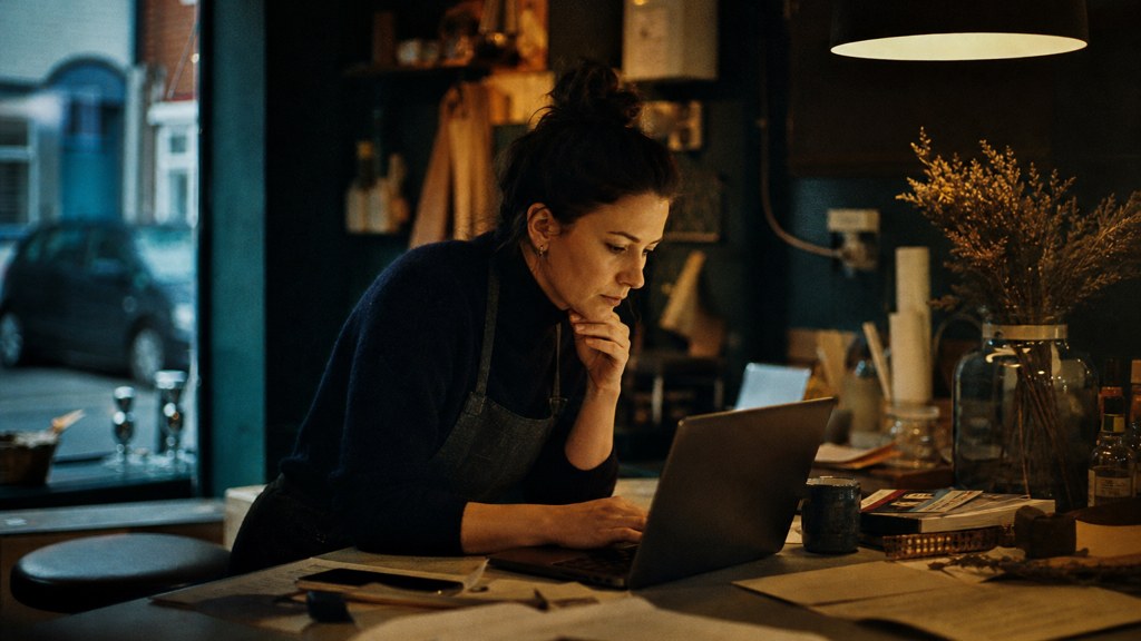 A small-business owner in an apron leaning over a laptop on a cluttered back-office desk late in the day, photographed in Dublin