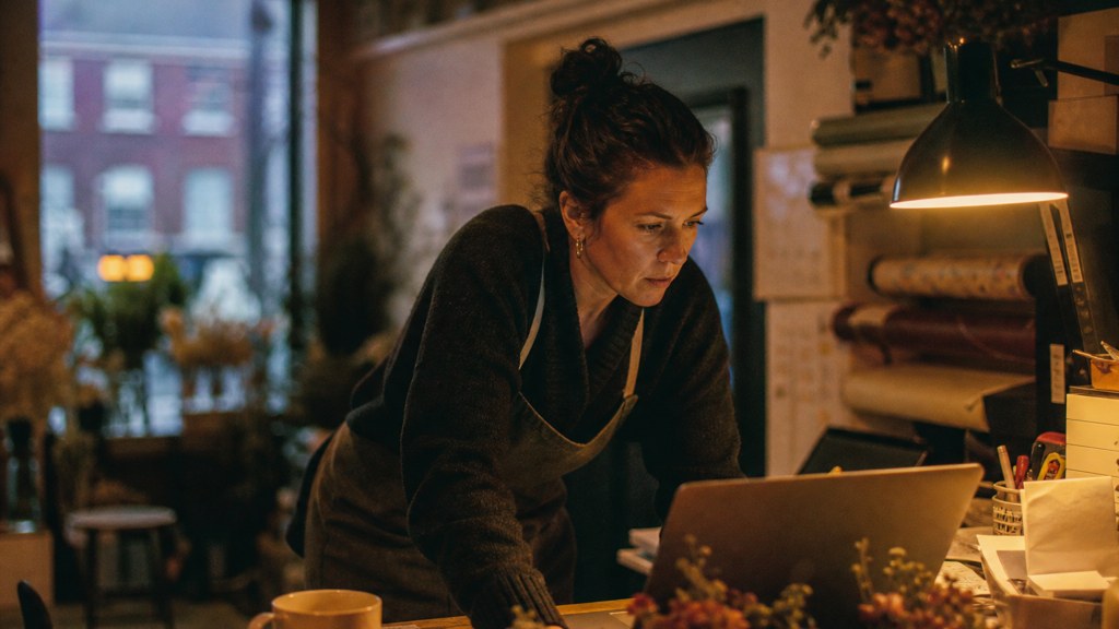 A small-business owner in an apron leaning over a laptop on a cluttered back-office desk late in the day, photographed in Dublin