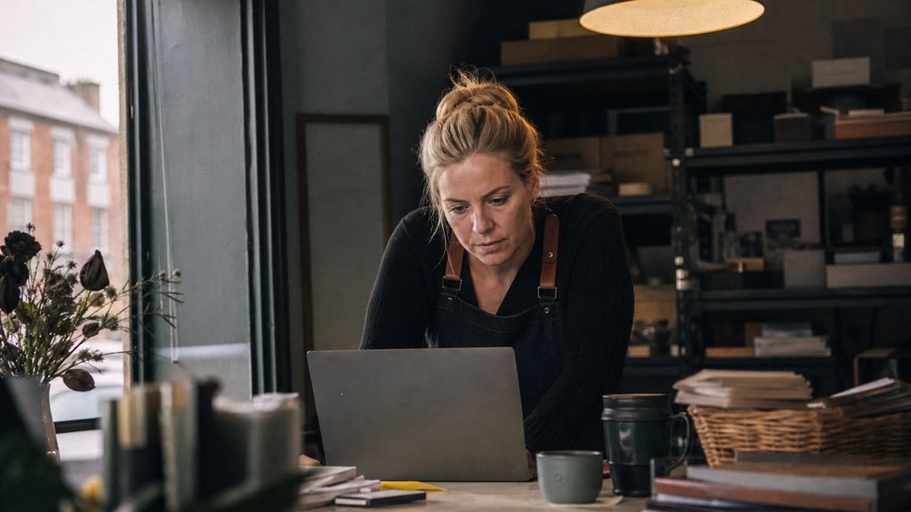 A small-business owner in an apron leaning over a laptop on a cluttered back-office desk late in the day, photographed in Dublin