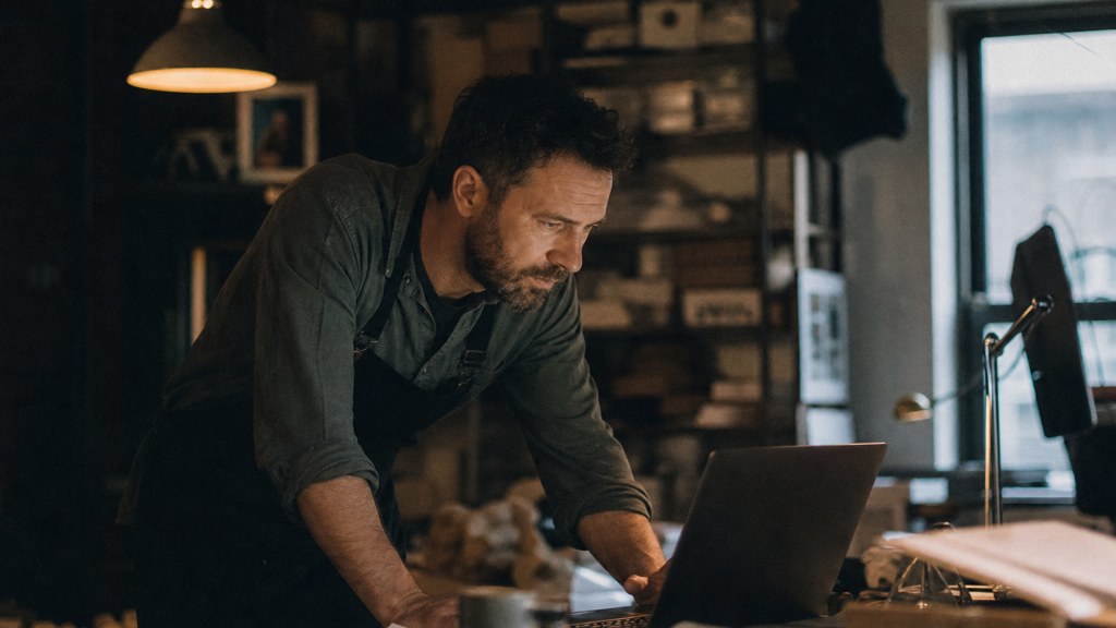 A small-business owner in an apron leaning over a laptop on a cluttered back-office desk late in the day, photographed in Durham