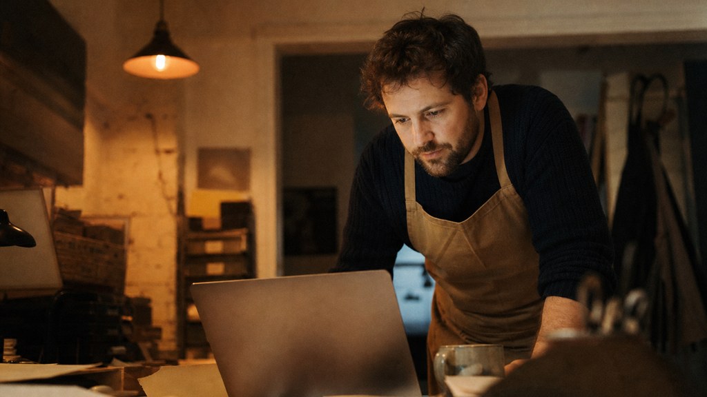 A small-business owner in an apron leaning over a laptop on a cluttered back-office desk late in the day, photographed in Durham