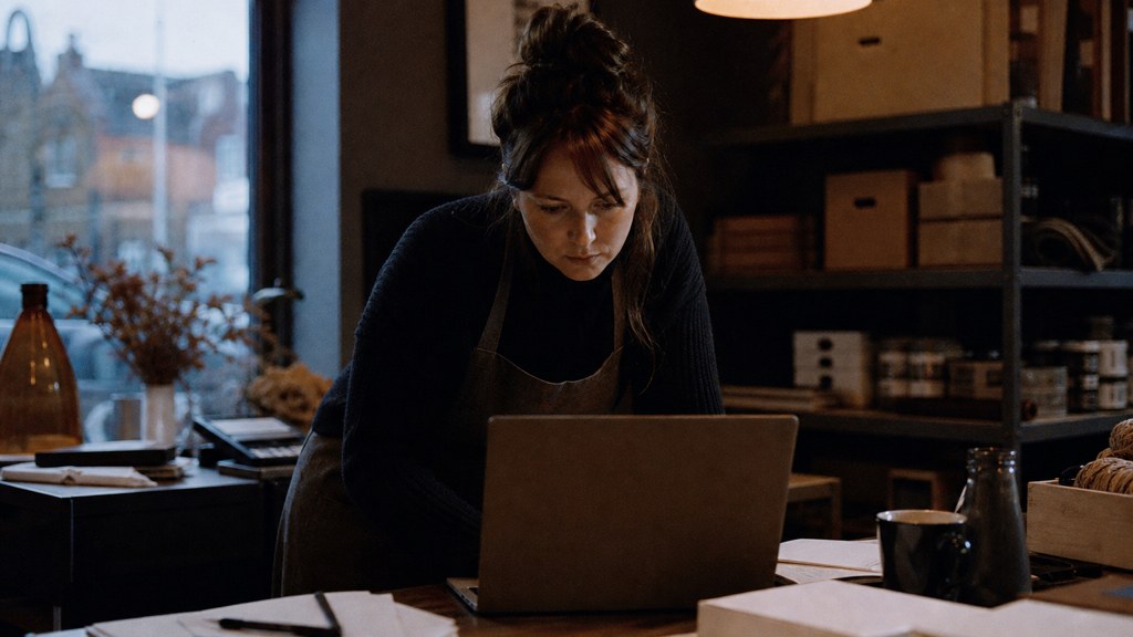 A small-business owner in an apron leaning over a laptop on a cluttered back-office desk late in the day, photographed in Durham